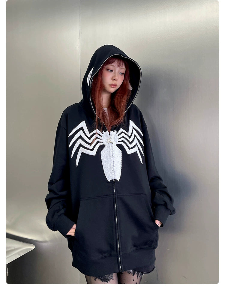 With long hair and confidence against a metallic wall, someone sports the Seakoff Venom Spider Hoodie—an oversized black and white zip-up sweatshirt. Perfect for streetwear fans, it features a bold white spider emblem on its 400GSM fabric.