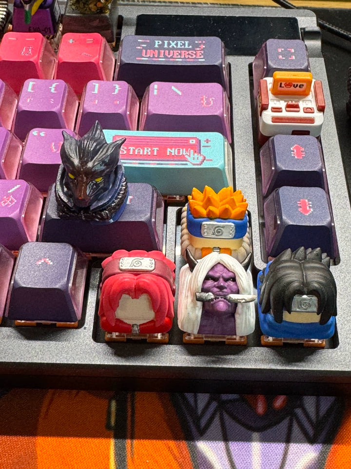A mechanical keyboard with colorful, themed keycaps. Several keycaps feature small, detailed character figures, each with distinctive hair and headbands. The keys have a pixelated design with labels like "Pixel Universe" and "Start Now.