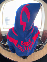 A person stands indoors by a window, face obscured, wearing the Seakoff Superhero Spider Hoodie—an oversized 400GSM blue and red zip-up sweatshirt for fans.