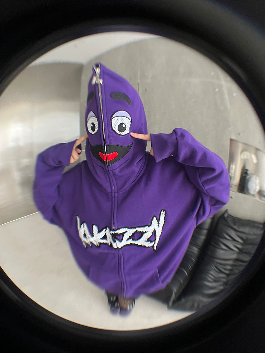 A person sports a Seakoff Fun Cartoon Face Hoodie, an oversized purple zip-up streetwear sweatshirt featuring a smiling face with eye and mouth cutouts. The fisheye lens effect blurs the room, enhancing the streetwear vibe.