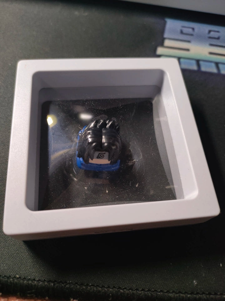A small black and blue keycap with a design resembling hair and a headband with a symbol is displayed inside a square, transparent case resting on a dark surface.
