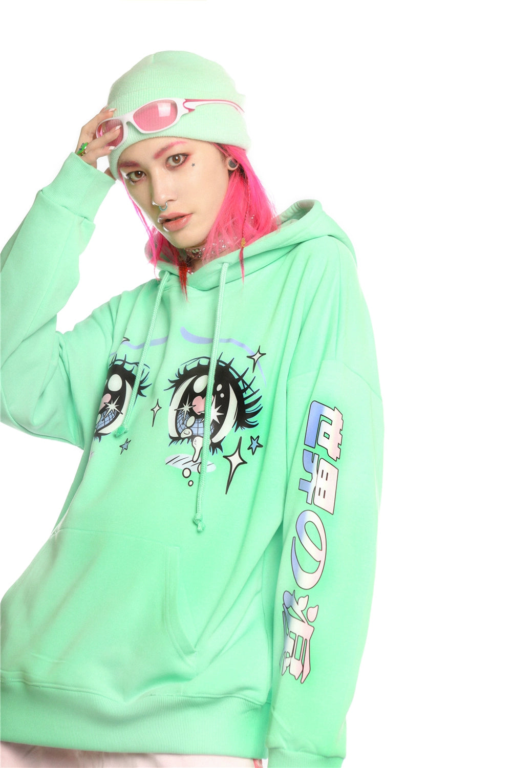 A person with pink hair showcases streetwear style in a Seakoff Kawaii Aesthetic Crying Eyes Hoodie – Pastel & Dark Anime Pullover featuring sleeve graphics. They complement the look with a matching beanie and pink goggles against a plain white backdrop.