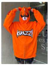 A person wears a Seakoff Furious Orange Bear Hoodie featuring an angry bear face, ears, and "KNAZZI" in bold. Made from premium 400GSM cotton with red embroidery partially covering their face, they stand before a concrete wall and window, enhancing the urban vibe.