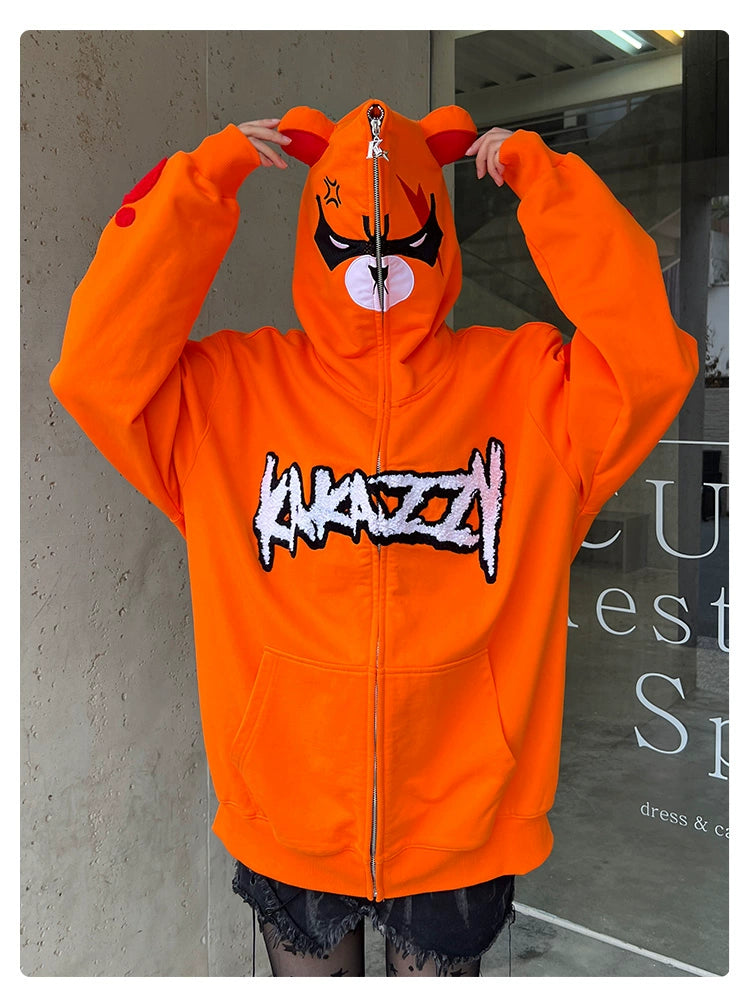 A person wears a Seakoff Furious Orange Bear Hoodie featuring an angry bear face, ears, and "KNAZZI" in bold. Made from premium 400GSM cotton with red embroidery partially covering their face, they stand before a concrete wall and window, enhancing the urban vibe.