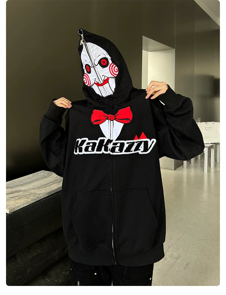 Someone wears a Seakoff Saw-Inspired Hoodie, an oversized black zip-up sweatshirt with a white mask design featuring red cheeks and spirals. The hoodie displays "Kakazy" in bold white. Perfect for horror movie fans, the outfit&