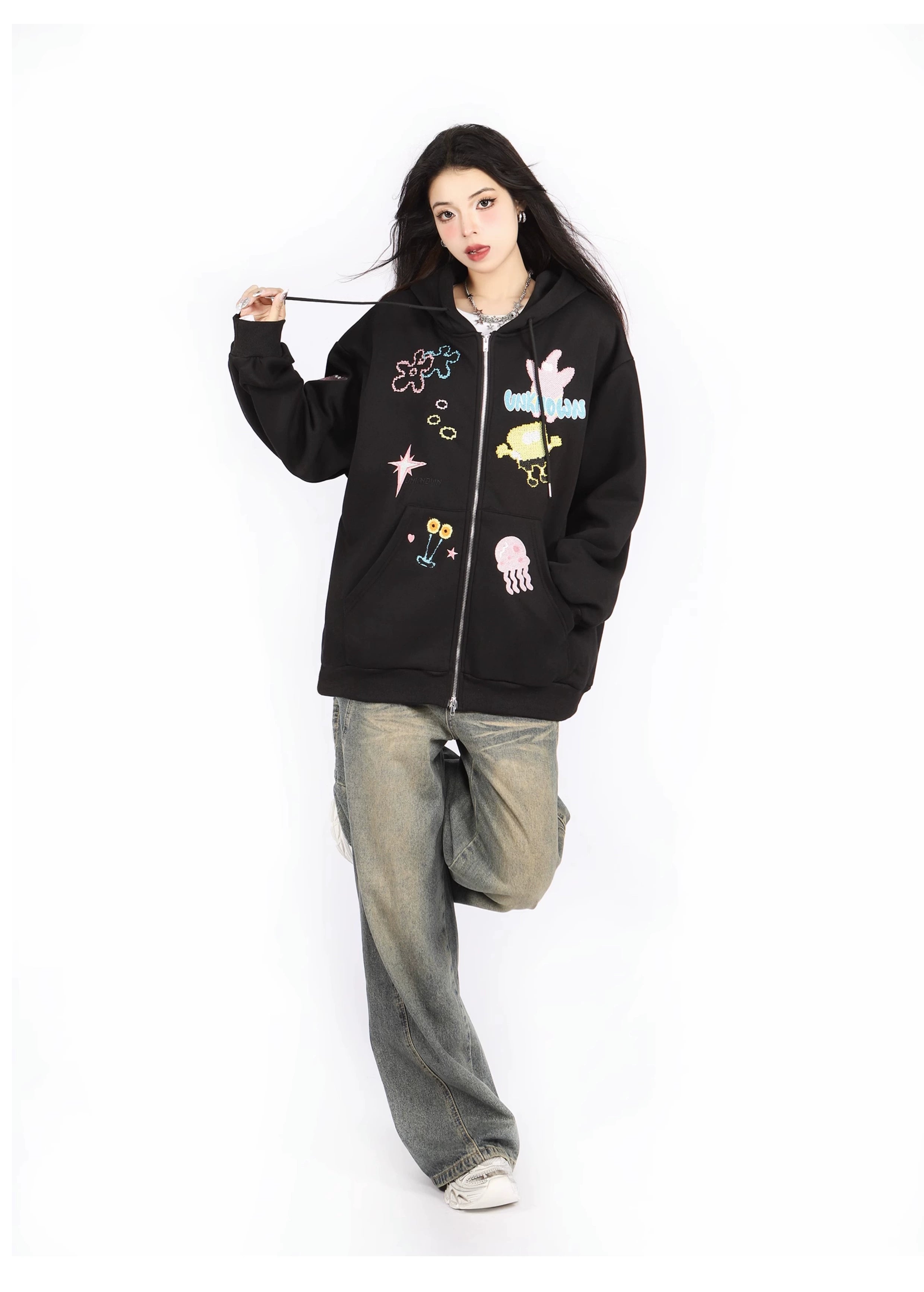 A person with long dark hair poses in a Seakoff Kawaii Pastel Cartoon Hoodie—featuring playful embroidered designs like jellyfish and spaceships. They embrace anime fashion with baggy jeans, white sneakers, and stand on one leg against a white background.