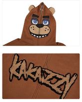 A close-up of the Seakoff Freddy-Inspired Hoodie shows a bear face with blue eyes, ears, nose, and mouth details. This oversized brown zip-up sweatshirt is ideal for horror game fans and features "KAKANZZY" in textured letters on the chest.