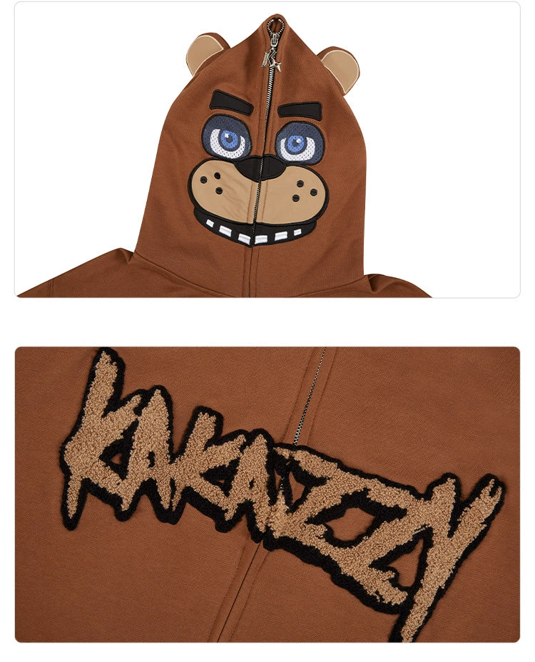 A close-up of the Seakoff Freddy-Inspired Hoodie shows a bear face with blue eyes, ears, nose, and mouth details. This oversized brown zip-up sweatshirt is ideal for horror game fans and features "KAKANZZY" in textured letters on the chest.