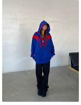 In a room, a person wears the Seakoff Superhero Spider Hoodie—an oversized blue and red zip-up sweatshirt perfect for fans. With the hood up, long hair flows over black pants and shoes. A black chair is partially visible to the right, enhancing the streetwear vibe.