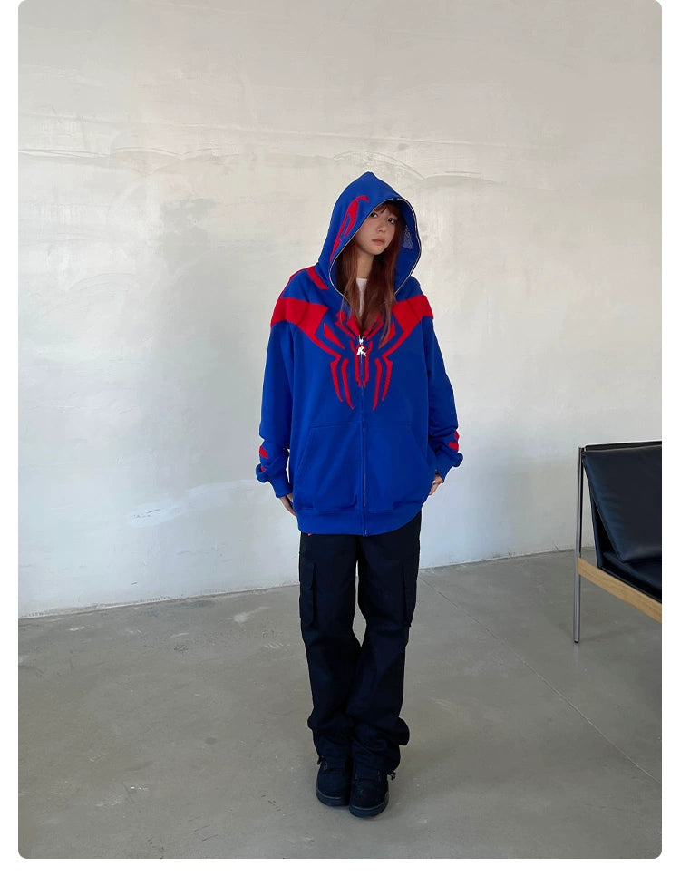 In a room, a person wears the Seakoff Superhero Spider Hoodie—an oversized blue and red zip-up sweatshirt perfect for fans. With the hood up, long hair flows over black pants and shoes. A black chair is partially visible to the right, enhancing the streetwear vibe.