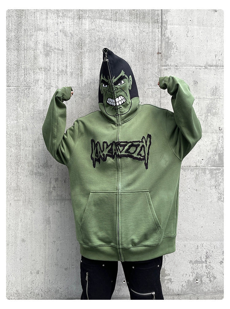 A person flexes in a Hulk-Inspired Hoodie by Seakoff, zipped up over their head with a menacing design and bold black lettering. This 400GSM oversized green superhero sweatshirt is perfect for fans and grabs the attention of streetwear enthusiasts against a concrete wall backdrop.