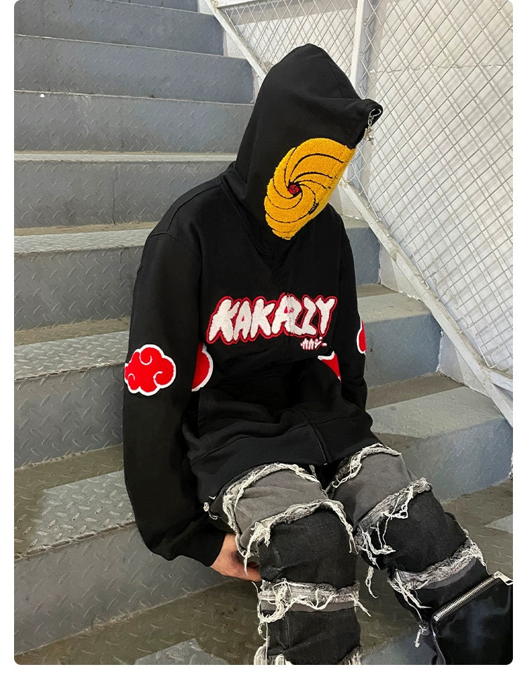 A person wearing a black Seakoff Uchiha Obito-Inspired Hoodie, featuring an Akatsuki Cloud Design and Spiral Mask, sits on a metal staircase. Perfect for Naruto fans, it has red cloud patches and "Kakazy" in red and white. They&