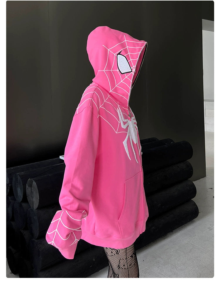 A person in a Seakoff Spider-Inspired Hoodie, 400GSM oversized pink zip-up sweatshirt with web design and large white spider, stands hidden by the hood. Superhero fans would love this against a dark stack of cylindrical objects and distant light.