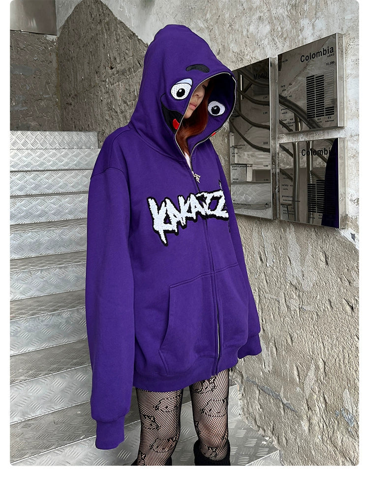 A person on stairs rocks streetwear with an oversized fit—a purple cartoon face hoodie from Seakoff, featuring eyes and an open mouth. "KINAZIC" is boldly imprinted on the front. Complemented by patterned fishnet tights, the scene is set against textured walls and mailboxes.