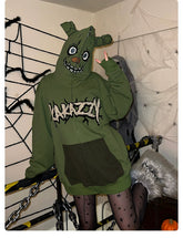 In a room decorated with spiderwebs and chains, someone wears a Seakoff Springtrap-Inspired Hoodie—a green oversized zip-up from Five Nights at Freddy&