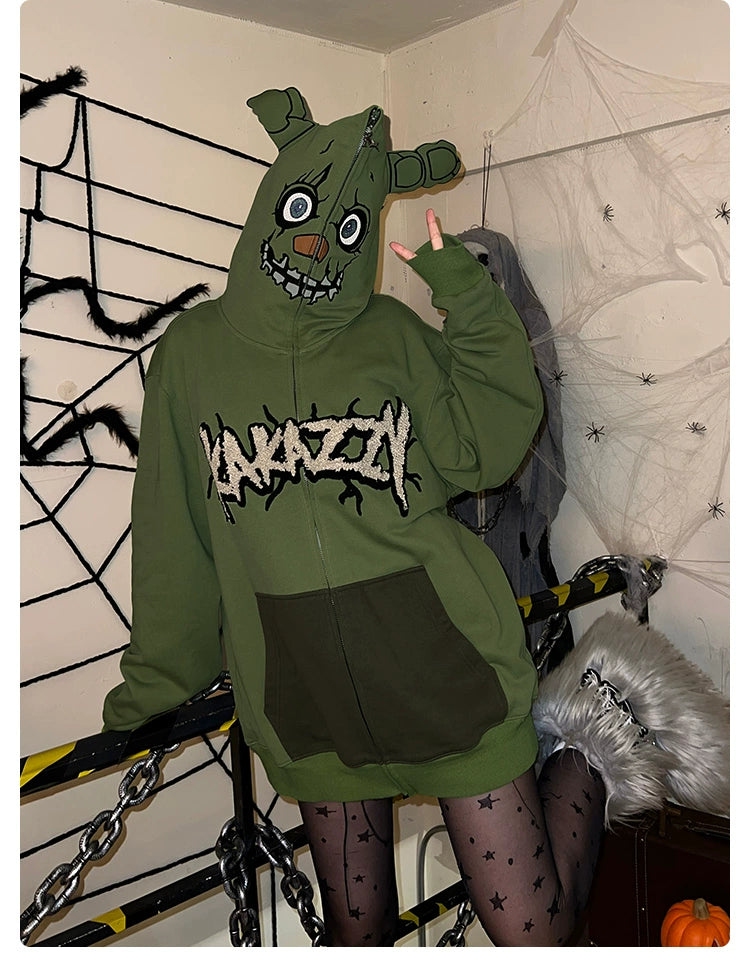 In a room decorated with spiderwebs and chains, someone wears a Seakoff Springtrap-Inspired Hoodie—a green oversized zip-up from Five Nights at Freddy&