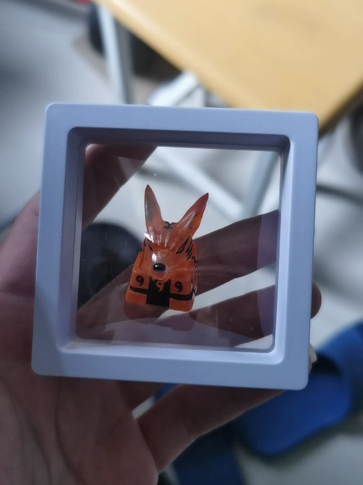A small orange fox figurine with the number "999" on it is encased in a white square display frame. The figure is held by a hand, and background details are slightly blurred, showing parts of a chair and table.