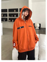 A person stands indoors in a Seakoff Fire Force Inspired Orange Zip-Up Hoodie, showcasing a character graphic and skull patch. The hood is up, blending with the minimalist decor of a white couch and wall shelves displaying small sculptures.