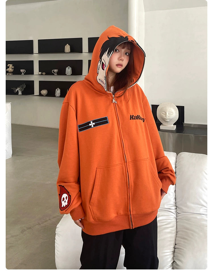 A person stands indoors in a Seakoff Fire Force Inspired Orange Zip-Up Hoodie, showcasing a character graphic and skull patch. The hood is up, blending with the minimalist decor of a white couch and wall shelves displaying small sculptures.