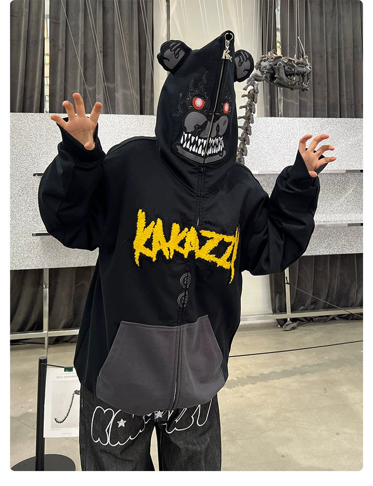 A person in the Seakoff Dark Bear Demon Hoodie, crafted from 400GSM cotton, raises their hands. The hoodie displays a scary bear face with hood ears and "KAZZZY" in yellow. Ideal for horror fans, their face is obscured against a textured backdrop with a skeleton lurking behind.