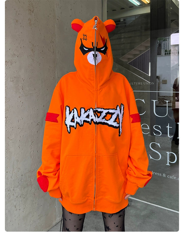 A person dons a Seakoff Furious Orange Bear Hoodie—an oversized 400GSM cotton piece featuring bear ears and an angry cartoonish bear face with closed eyes. "KANAZZY" is printed on the chest. The scene includes a glass door and concrete wall backdrop.