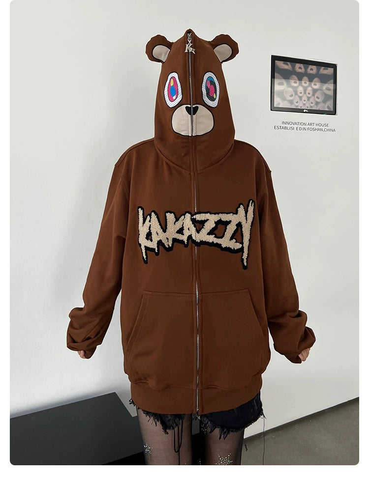 The person is sporting a Seakoff Cute Bear Hoodie, an oversized 400GSM brown zip-up sweatshirt featuring a teddy bear face with ears and colorful eyes on the hood. The furry-textured "KUNAZY" text adorns the front, completing the streetwear look with hands coolly in pockets.