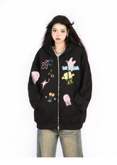 Against a white backdrop, someone wears the Seakoff Kawaii Pastel Cartoon Hoodie, showcasing playful embroidered designs of jellyfish, flowers, and abstract shapes that echo anime fashion. This oversized black hoodie perfectly complements loose-fitting jeans for a playful yet stylish look.