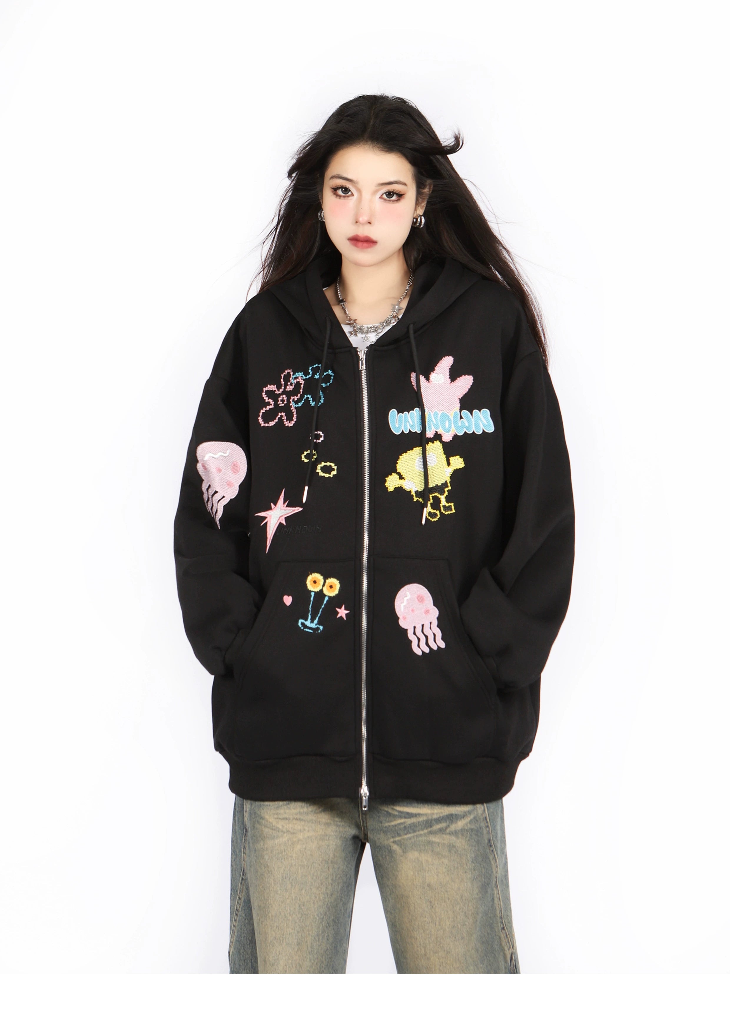 Against a white backdrop, someone wears the Seakoff Kawaii Pastel Cartoon Hoodie, showcasing playful embroidered designs of jellyfish, flowers, and abstract shapes that echo anime fashion. This oversized black hoodie perfectly complements loose-fitting jeans for a playful yet stylish look.