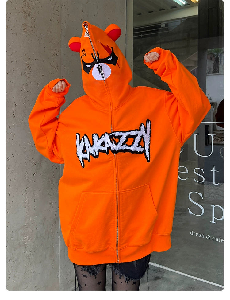 Someone is wearing a Seakoff Furious Orange Bear Hoodie made of premium 400GSM cotton. It features an oversized design with an angry bear face on the hood and "CRAZY" printed on the front. Raising their fists, they zip up the hood to cover their face.