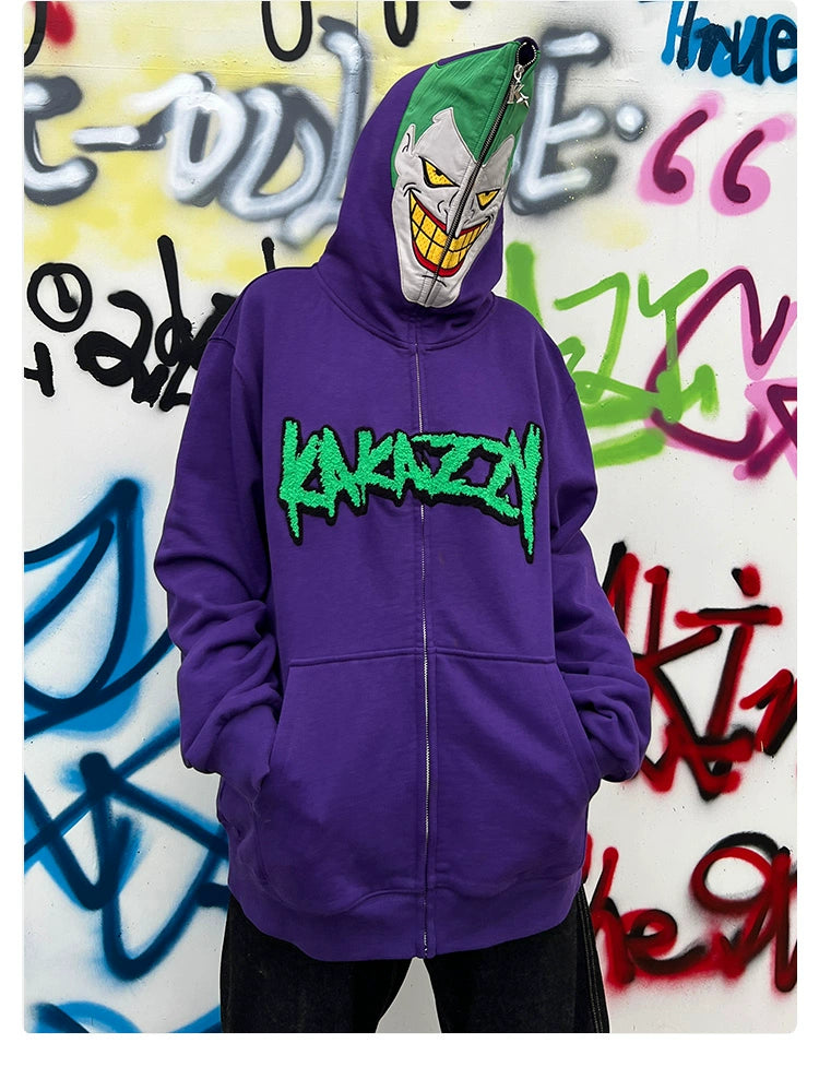 A person dons a Seakoff Joker-Inspired Hoodie, an oversized 400GSM cotton purple zip-up sweatshirt, featuring "Kakazzy" in green. With a masked face evoking a cartoonish grin and green hair top, they pose against graffiti—ideal for supervillain enthusiasts.