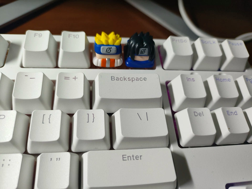 A white keyboard features two keycaps designed as characters with spiky yellow and black hair and headbands. The larger "Backspace" key is prominently displayed in the center.