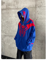A person stands against a concrete wall in a Seakoff Superhero Spider Hoodie, an oversized 400GSM blue and red zip-up sweatshirt with a striking spider emblem. Their hands are tucked in the pockets, and the hood is raised, partially concealing their face.