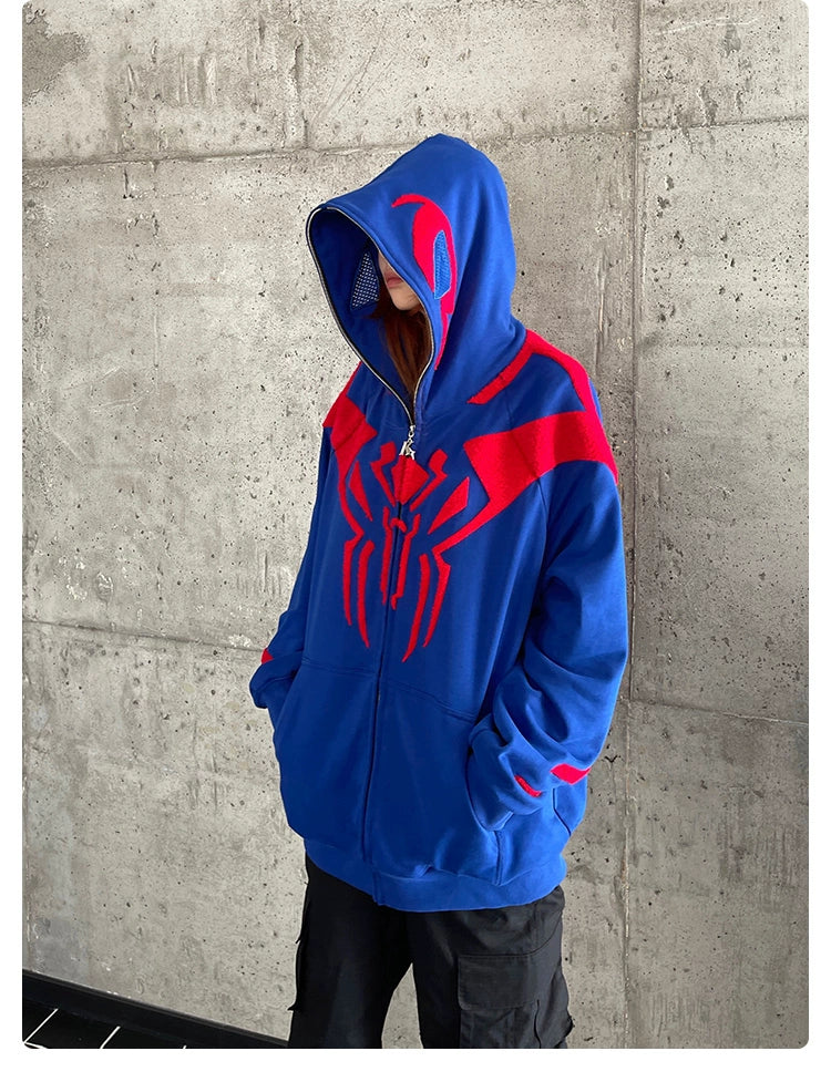 A person stands against a concrete wall in a Seakoff Superhero Spider Hoodie, an oversized 400GSM blue and red zip-up sweatshirt with a striking spider emblem. Their hands are tucked in the pockets, and the hood is raised, partially concealing their face.