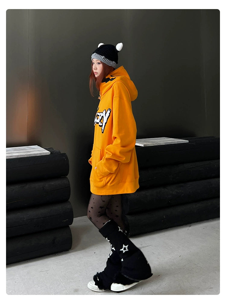 A person is indoors wearing a vibrant Seakoff Halloween Pumpkin Hoodie, star-patterned black leggings, and black boots, paired with a black-and-white beanie with pom-poms. A stack of large cushions sits beside them, enhancing the festive ambiance against the dark background.