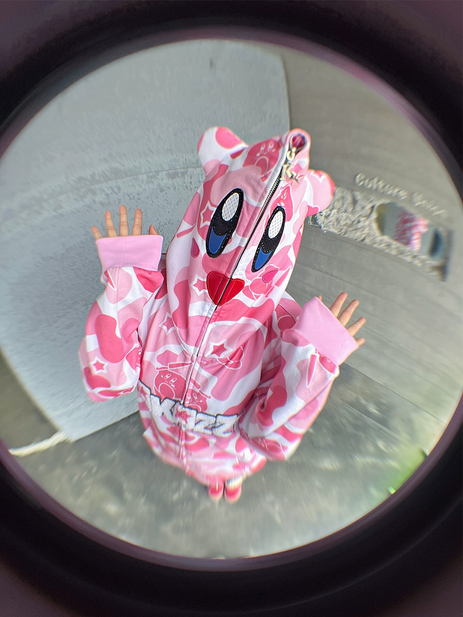 A person in a Seakoff Pink Kirby-Inspired Hoodie with camo design and 3D ears poses with raised arms. The hoodie, made of 400GSM cotton, features large eyes and a wide mouth, enhanced by the fisheye lens for extra whimsy.