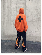 A person faces away from the camera in a Seakoff Fire Force Inspired Orange Zip-Up Hoodie, featuring a black cross design. They style it with black pants accented with orange and matching shoes, creating an anime streetwear look against a plain concrete wall.