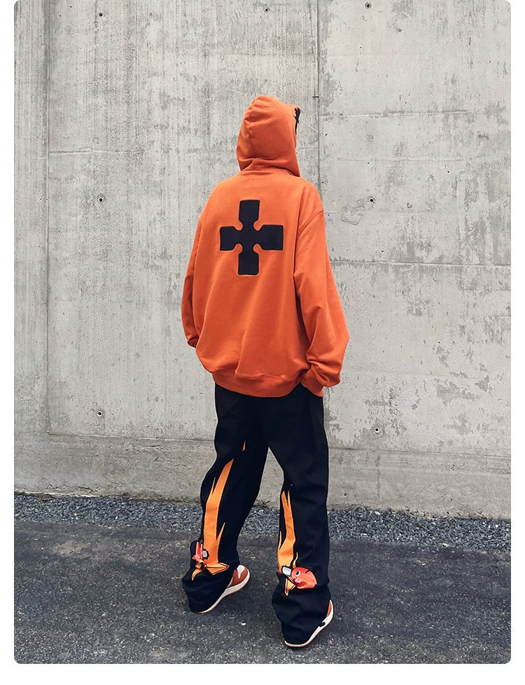 A person faces away from the camera in a Seakoff Fire Force Inspired Orange Zip-Up Hoodie, featuring a black cross design. They style it with black pants accented with orange and matching shoes, creating an anime streetwear look against a plain concrete wall.