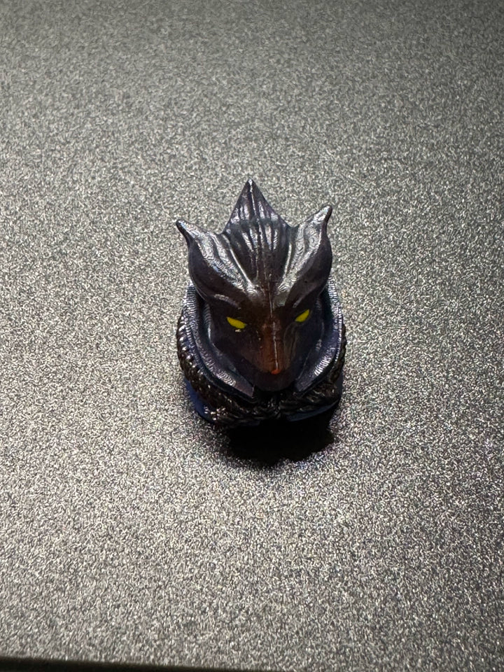 A small, sculpted figure resembling a mythical creature with pointed ears, glowing yellow eyes, and a textured surface is placed on a gray, textured background.