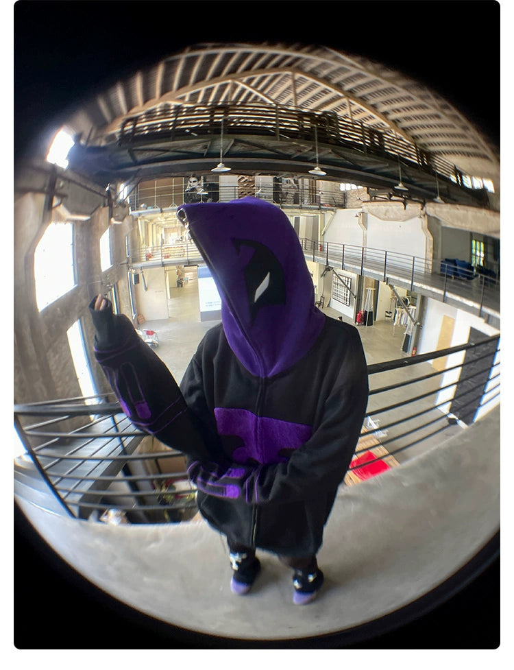 A person stands inside on a balcony, wearing the Seakoff Dark Hero Hoodie, an oversized black and purple superhero zip-up sweatshirt. The hood is up, concealing their face. Captured with a fisheye lens, the image has a unique curved effect, set against an interior with exposed beams.