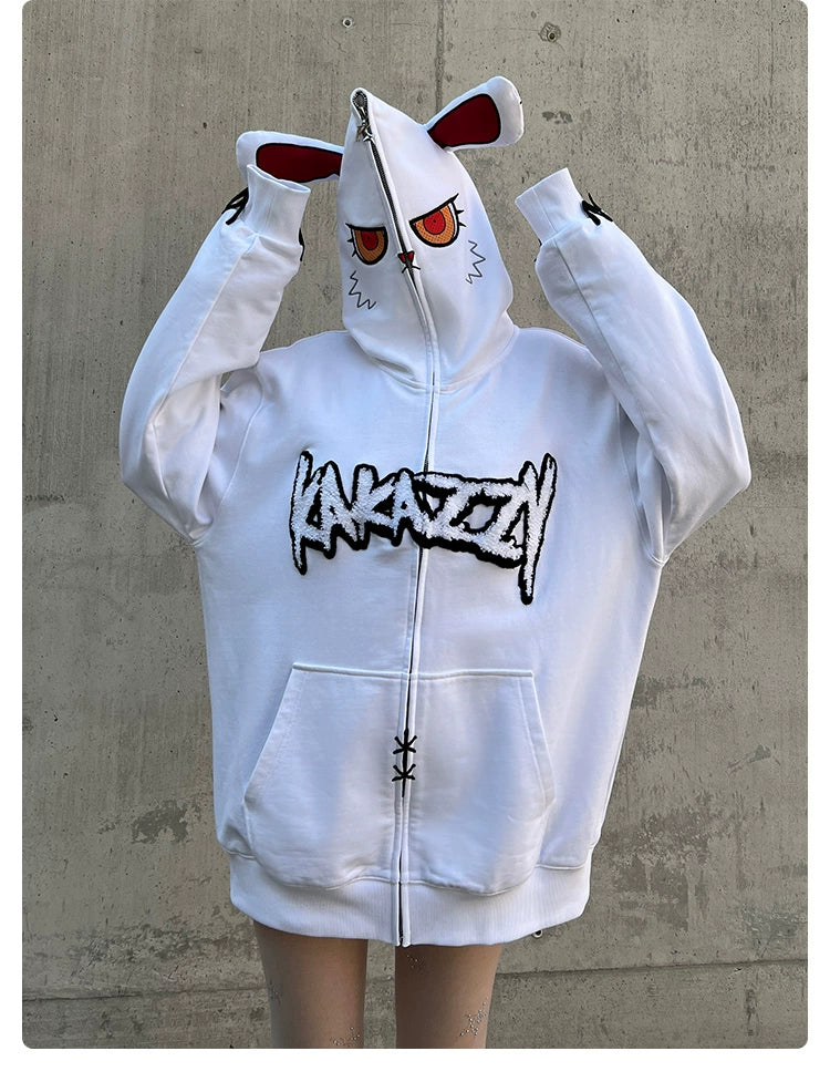 A person in a Seakoff Fierce Bunny Hoodie, showcasing an oversized fit with rabbit ears and cartoonish red eyes on the hood. The front reads "KARAZZI" in bold black text as the hood is zipped up partially covering their face while they stand against a concrete wall.