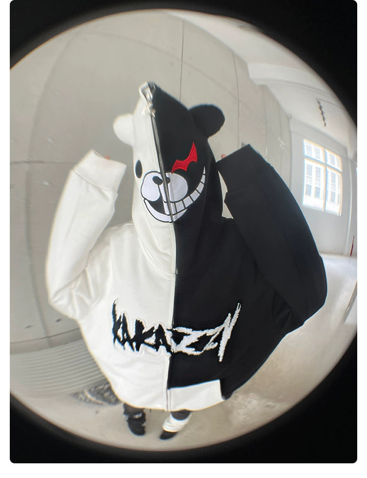 A Danganronpa fan wearing a Seakoff Monokuma-Inspired Hoodie, featuring a mischievous smile and red eye on the black half and "MAZZY" in textured letters, stands in a light gray room.