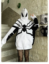 Standing on stairs making a peace sign, a person wears the Seakoff Venom-Inspired White Hoodie, an oversized zip-up superhero sweatshirt featuring spider web and eye designs made from 400GSM cotton. Star-patterned tights add flair against textured concrete walls adorned with posters.