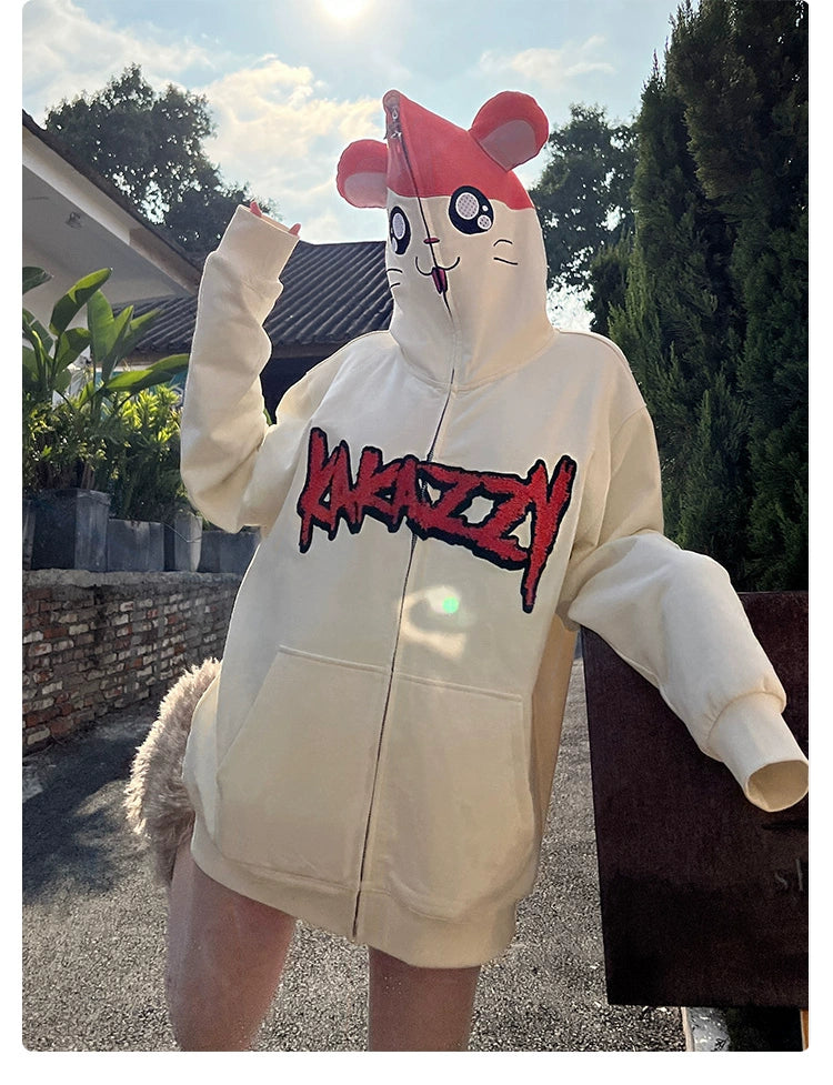 A person outdoors wears a Seakoff Cute Hamster Cartoon Hoodie made of 400GSM cotton, featuring playful large pink ears and "ANAZAZY" in red on the front. They stand on a paved path with greenery and a building in the background, making this hoodie an ideal gift for cartoon lovers.