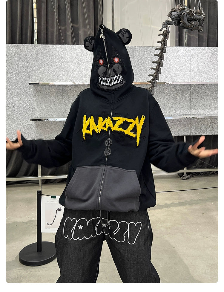 A person wears the Seakoff Dark Bear Demon Hoodie, 400GSM cotton with a bear mask featuring a zippered mouth and red eyes. "Kakazzy" marks the black hoodie and pants against a gray wall with a dinosaur skeleton display, ideal for horror fans.
