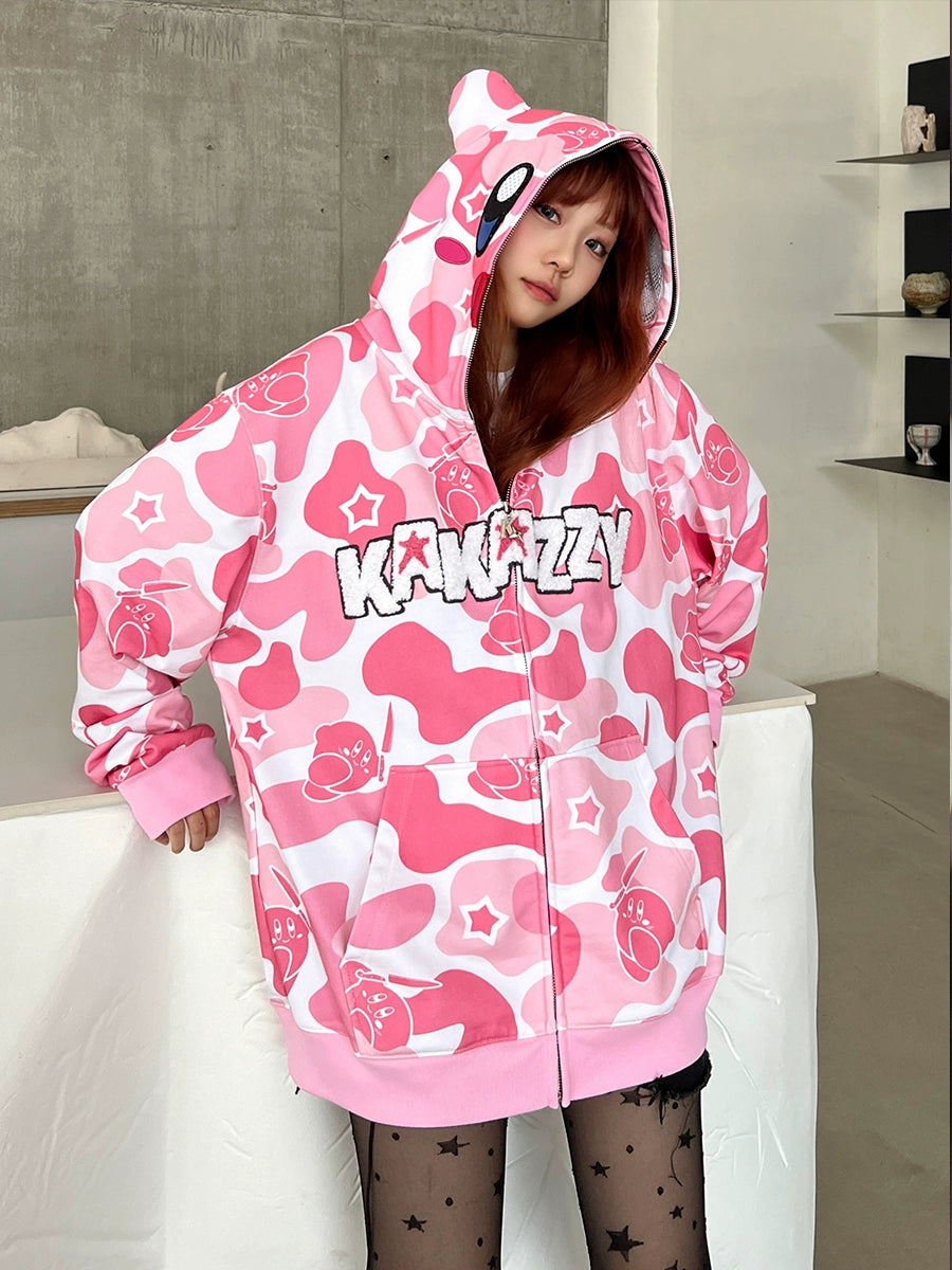 A person wearing the Seakoff Pink Kirby-Inspired Hoodie, featuring a pink and white camo design, cartoon graphics, and "Kawaii" text with charming 3D ears. They lean indoors against a white surface with a neutral expression, set against a concrete wall and shelves.