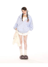 A person embraces kawaii fashion against a plain white background, wearing a Seakoff Hello Kitty Pink Kawaii Hoodie with cat ear hood, white textured leggings, and fluffy slippers. They hold a fluffy white bag and sport pigtails.