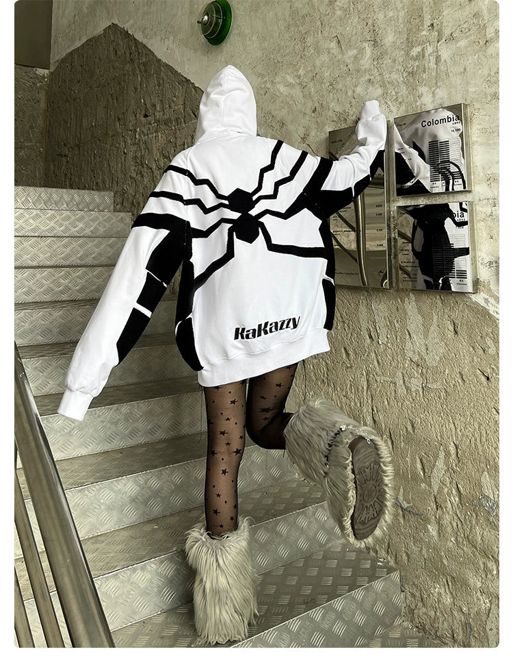 Wearing a Seakoff Venom-Inspired White Hoodie, featuring a black spider emblem and "Rakazzy" text, a person ascends stairs. Made of 400GSM cotton for a premium feel, they pair it with furry boots and patterned tights. Reflective surfaces and textured concrete walls complete the scene.