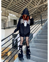A person in a Seakoff Venom Spider Hoodie, an oversized 400GSM black and white zip-up sweatshirt for superhero fans, stands on metal stairs. Their red hair contrasts with star-patterned tights and leg warmers, turning heads among streetwear enthusiasts amid metal railings and high ceilings.