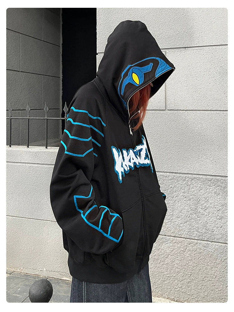 Wearing a Seakoff Cyberpunk Superhero-Inspired Hoodie, an oversized 400GSM black zip-up sweatshirt with a blue "KRAKEN" design, they stand hooded against a textured gray wall and spiked fence—perfect for sci-fi fans embracing futuristic styles.