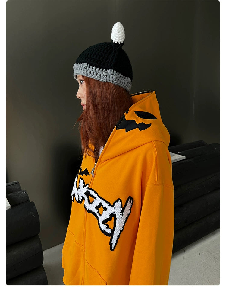A person with long brown hair wears a black and white knitted hat and an orange Seakoff Halloween Pumpkin Hoodie, a 400GSM oversized zip-up for fans, with bold black and white lettering. The dark gray background has vague black objects, adding to the mysterious vibe.